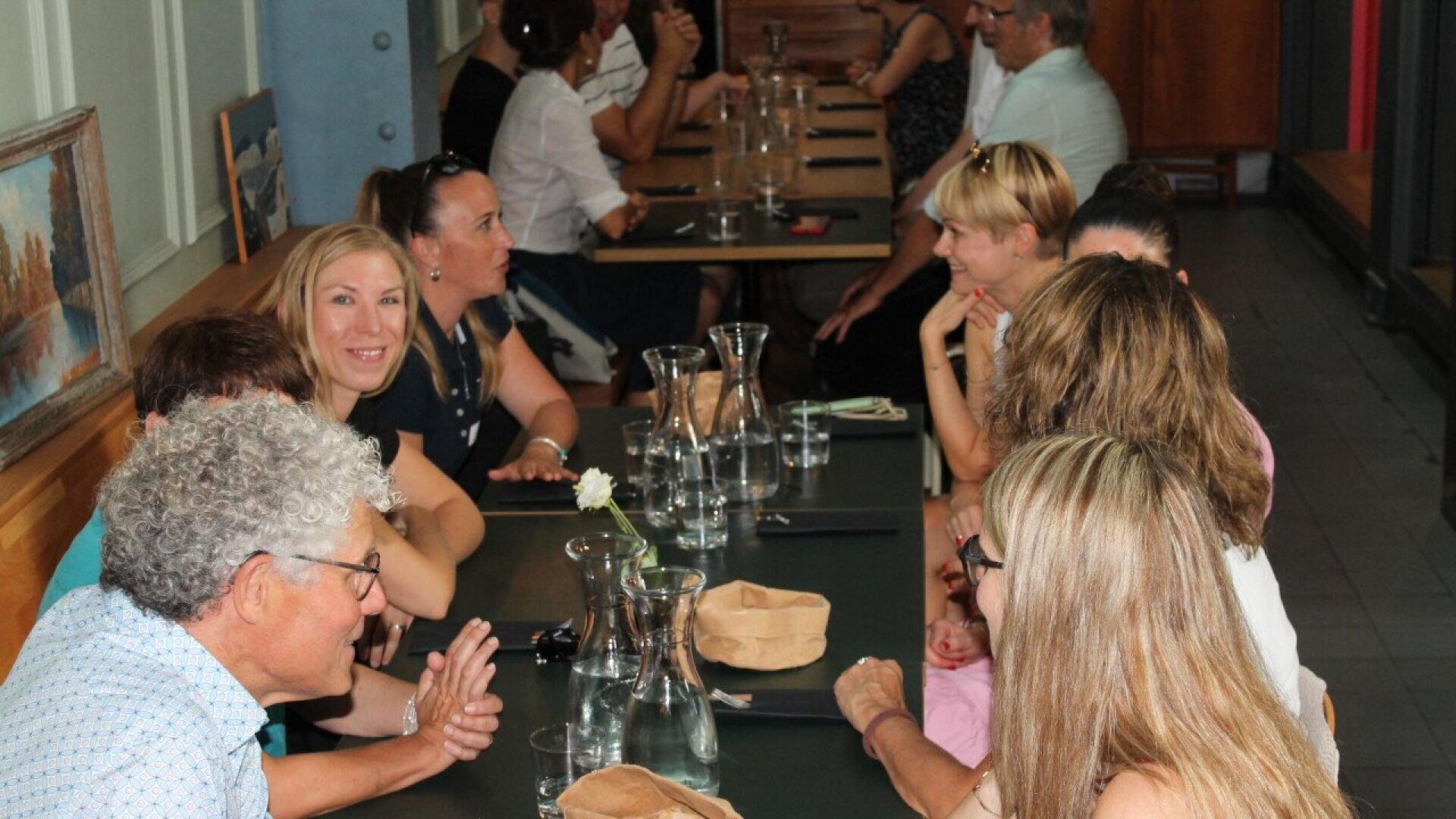 A group of people sit at a long table in a restaurant, engaged in conversation and smiling, with glass pitchers and brown paper bags on the table. The setting is casual and lively.