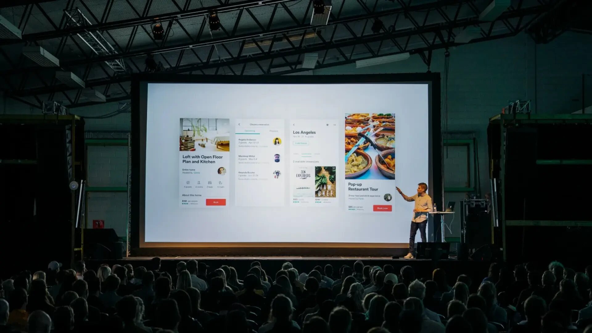 A person stands on stage presenting to a large seated audience, with a giant screen displaying a user interface featuring cards for locations, people, and restaurants.