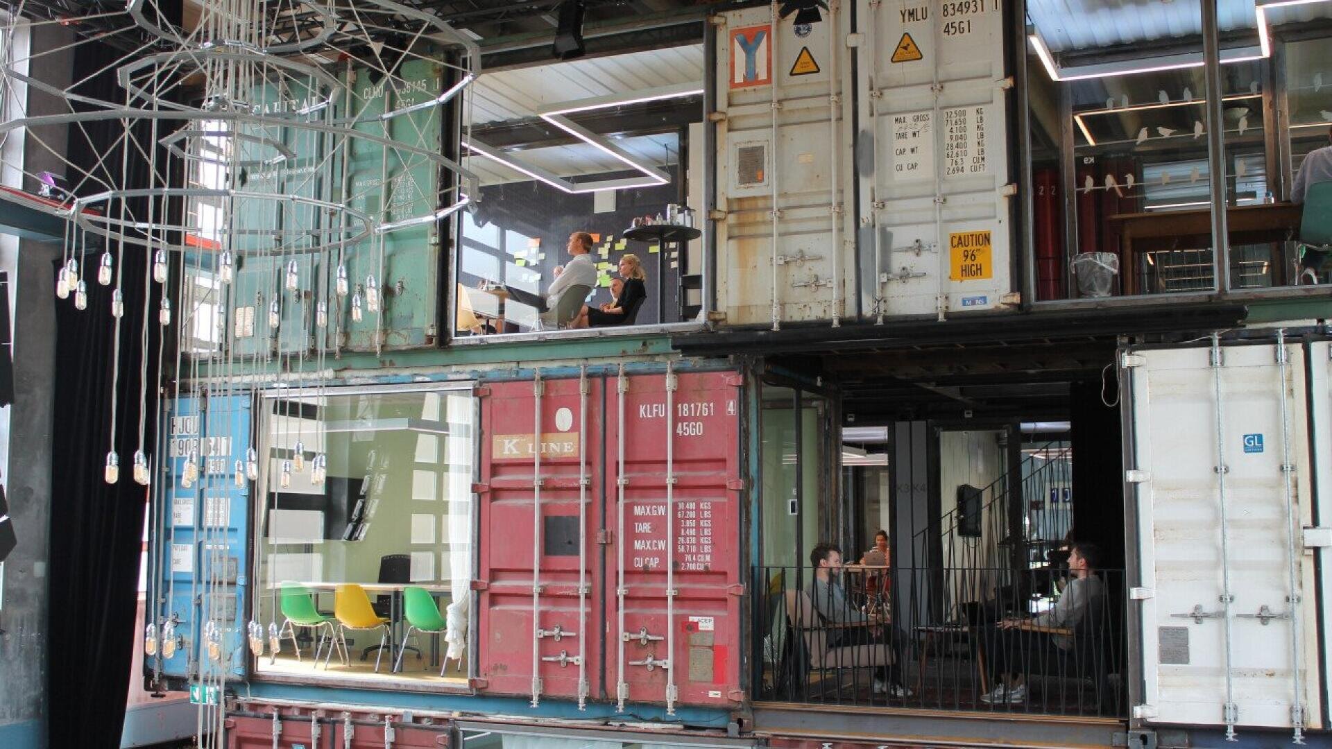 Two stacked shipping containers repurposed as offices and meeting spaces, with people working inside. Colorful chairs, modern decor, and wires hanging in the foreground create an industrial yet creative atmosphere.