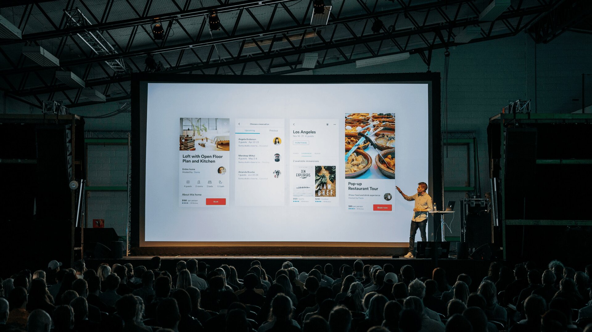 Eine Person steht auf der Bühne und hält vor einem großen Publikum eine Präsentation. Auf einem riesigen Bildschirm werden App-Oberflächendesigns mit den Themen Essen und Reisen angezeigt.