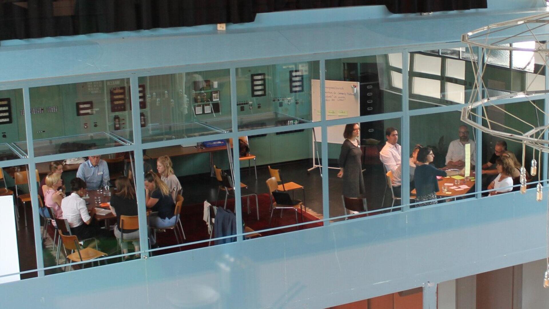 A group of people sit around tables in a modern glass-walled room, engaged in discussion and activities, while a few others stand and talk near a whiteboard in the background.