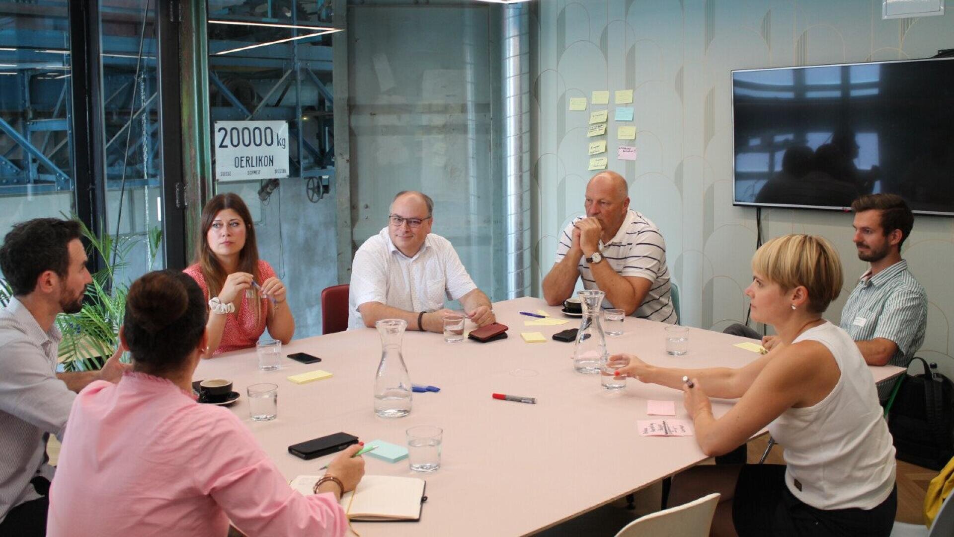 Seven people sit around a conference table engaged in discussion, with notebooks, pens, and glasses of water in front of them. Sticky notes are on the wall, and a large TV screen is mounted nearby.
