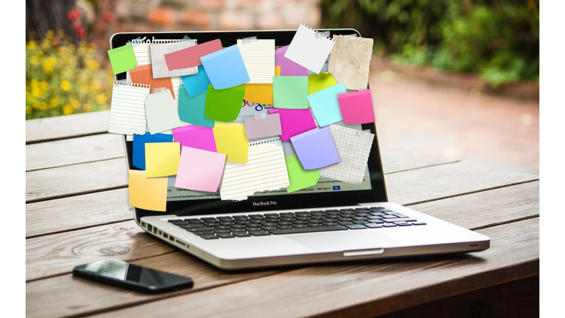 Ein Laptop, der mit bunten Haftnotizen und Papier bedeckt ist und den Bildschirm verdeckt, steht auf einem Holztisch neben einem Smartphone und einem Notebook und lässt einen geschäftigen oder überfüllten Arbeitsplatz vermuten.