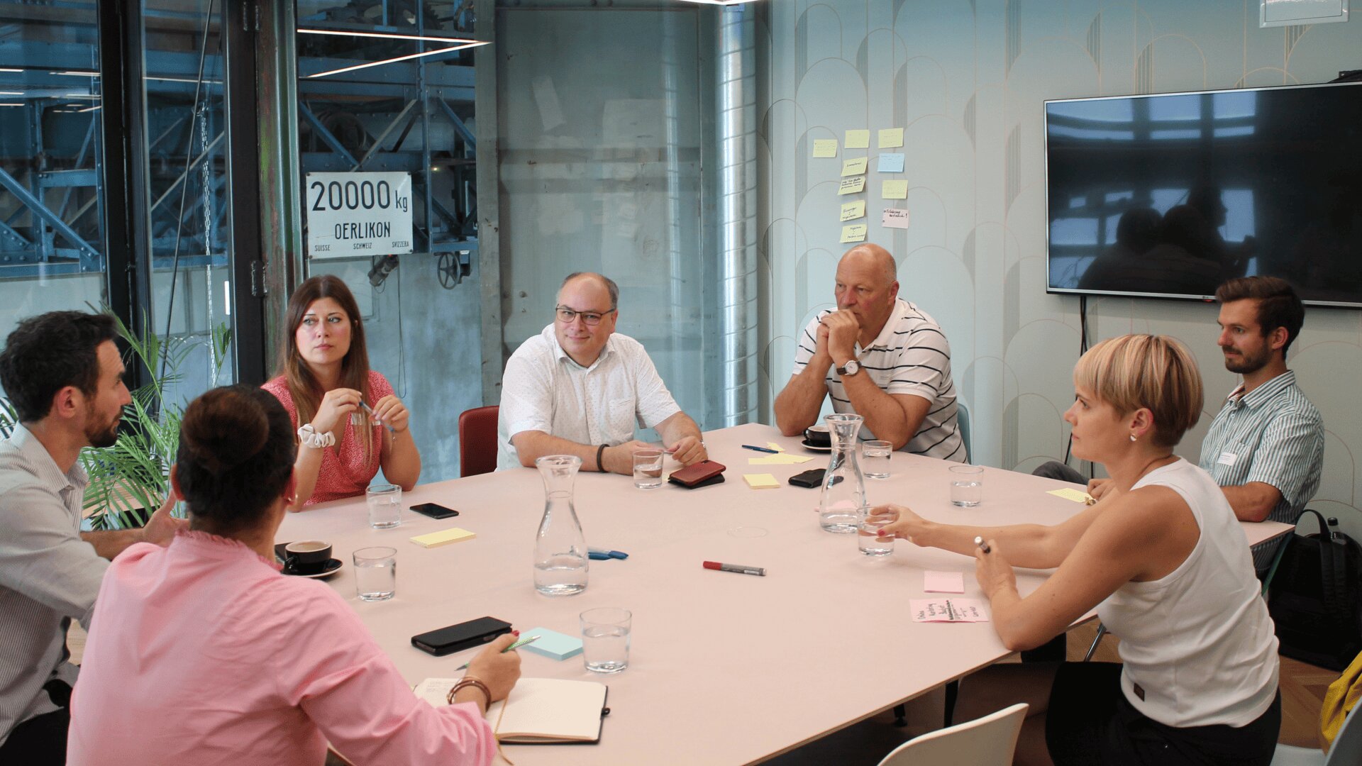 Sieben Personen sitzen um einen Konferenztisch und diskutieren. Notizbücher, Stifte und Wassergläser stehen auf dem Tisch. An der Wand hängen Haftnotizen, im Hintergrund ist ein großer Bildschirm zu sehen.