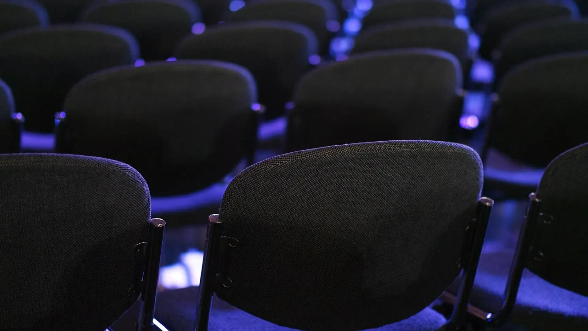 Rows of empty, dark-colored chairs are arranged in an auditorium or conference setting, with soft blue lighting illuminating the scene.