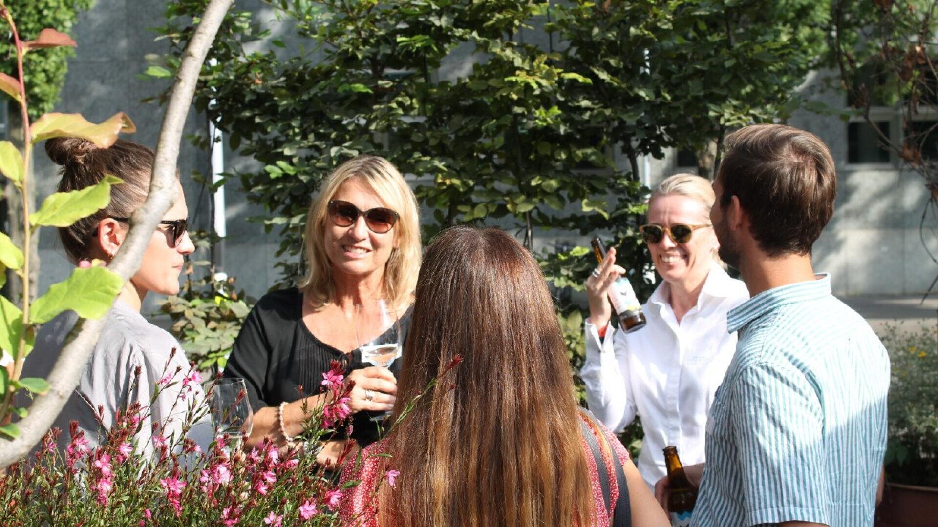A group of five adults stand outside in a garden, talking and smiling while holding drinks. Sunlight filters through the trees, creating a bright, relaxed atmosphere. Some people wear sunglasses.