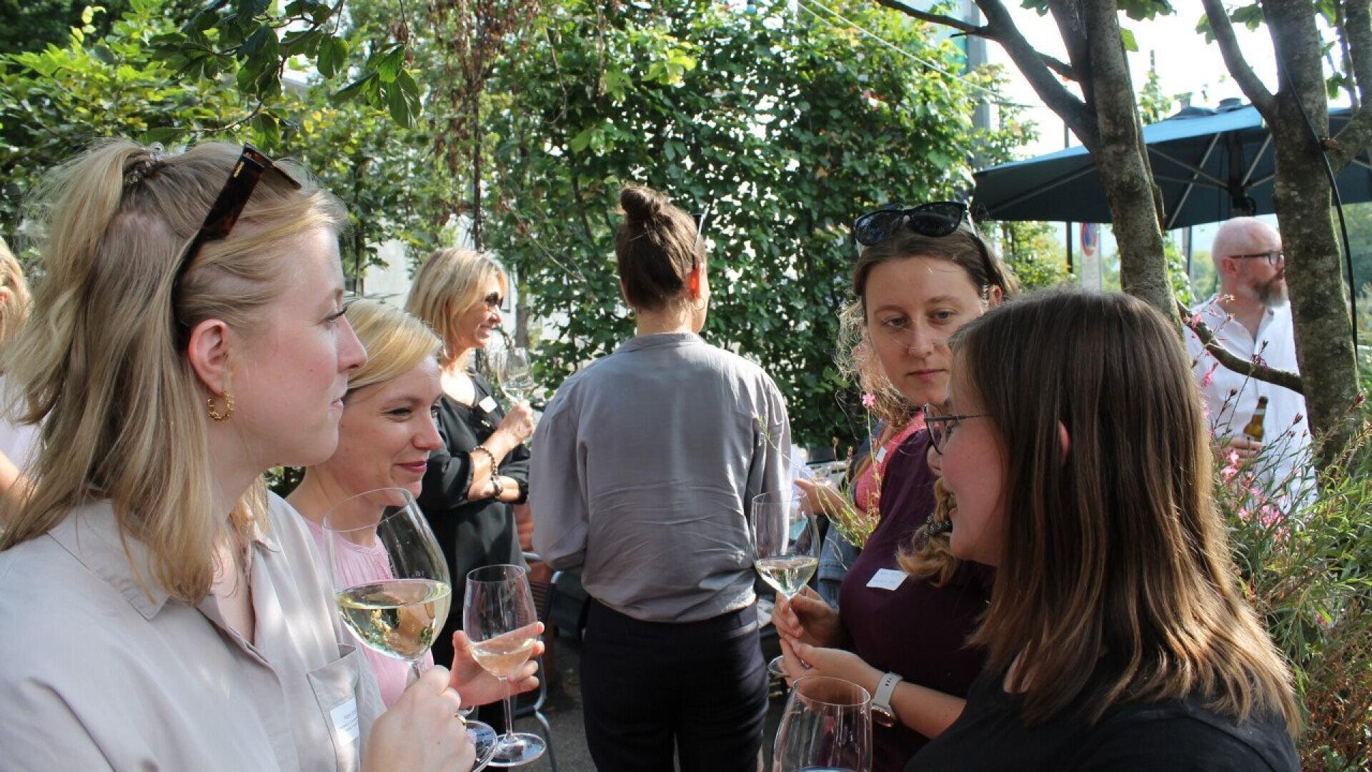 A group of people stand outdoors, holding glasses and talking in a garden setting. Trees and greenery surround them, and most appear engaged in conversation, enjoying a social gathering.