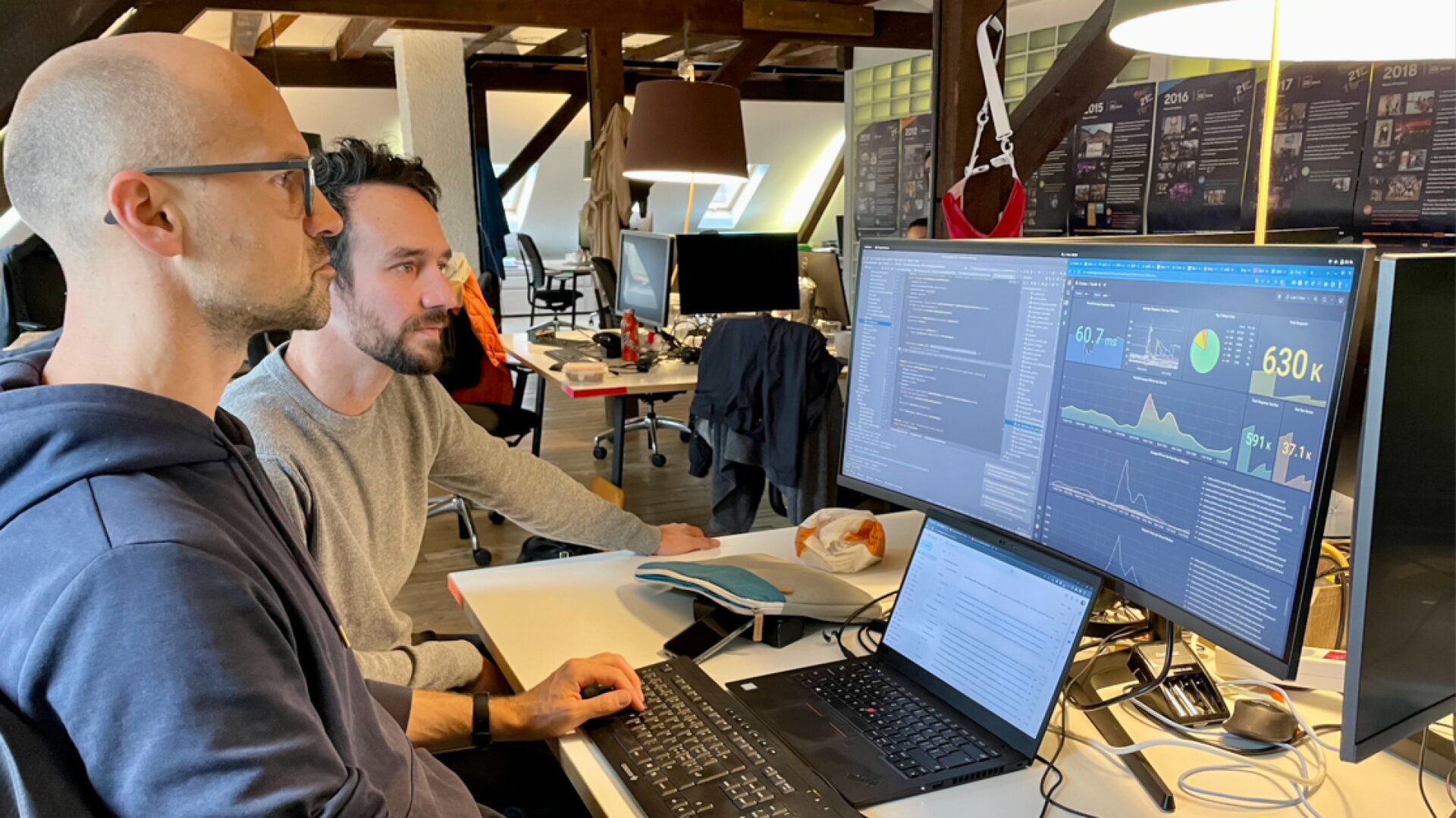 Zwei Männer sitzen an einem Schreibtisch in einem modernen Büro und sehen sich Datenvisualisierungen und Codes auf zwei Monitoren genau an. Der Arbeitsbereich besteht aus freiliegenden Balken, Laptops und Büromaterialien, die überall verstreut sind.