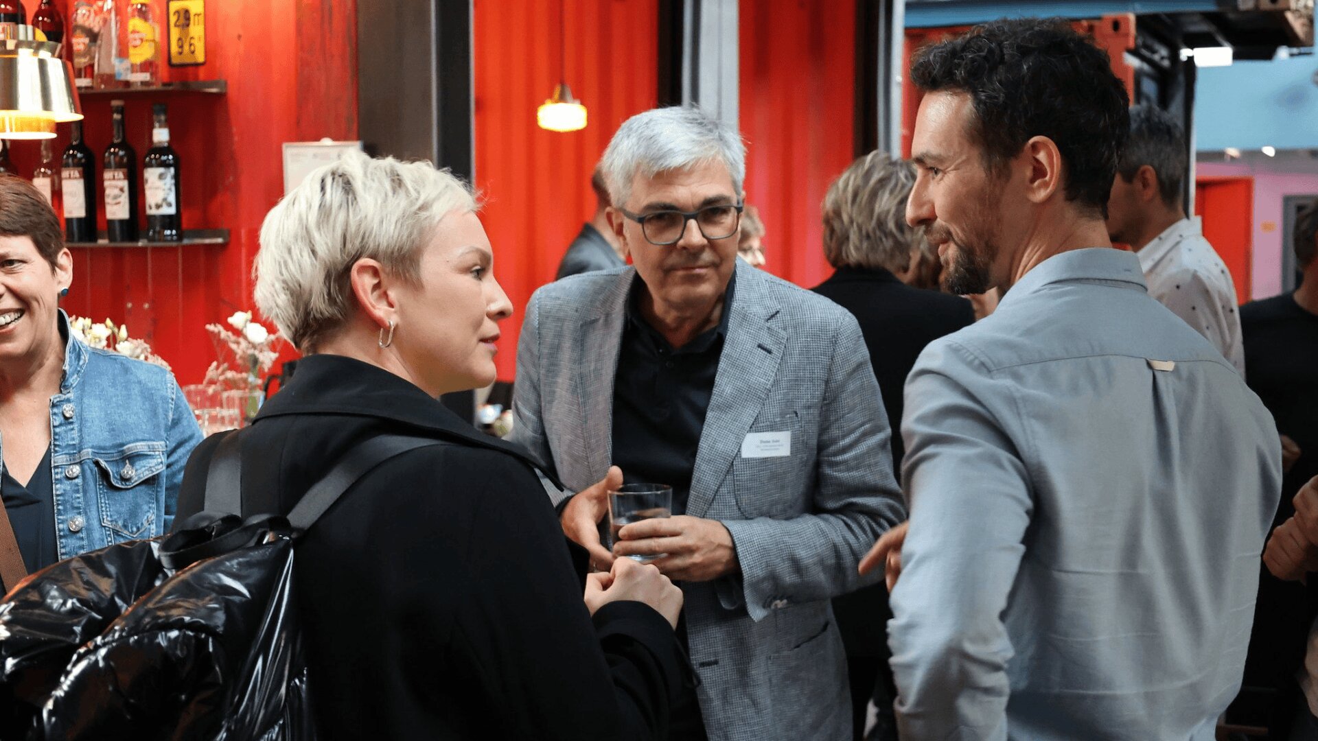 Eine Gruppe von Menschen trifft sich in einem Innenbereich, der wie eine zwanglose Networking-Veranstaltung aussieht. Einige unterhalten sich, einer hält ein Getränk in der Hand und auf Regalen vor einem roten Hintergrund sind Flaschen zu sehen.
