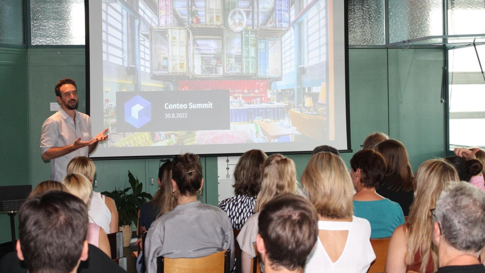A man stands in front of an audience giving a presentation at the Confeo Summit. A large screen behind him displays Confeo Summit 30.8.2022 with colorful graphics and architectural images.