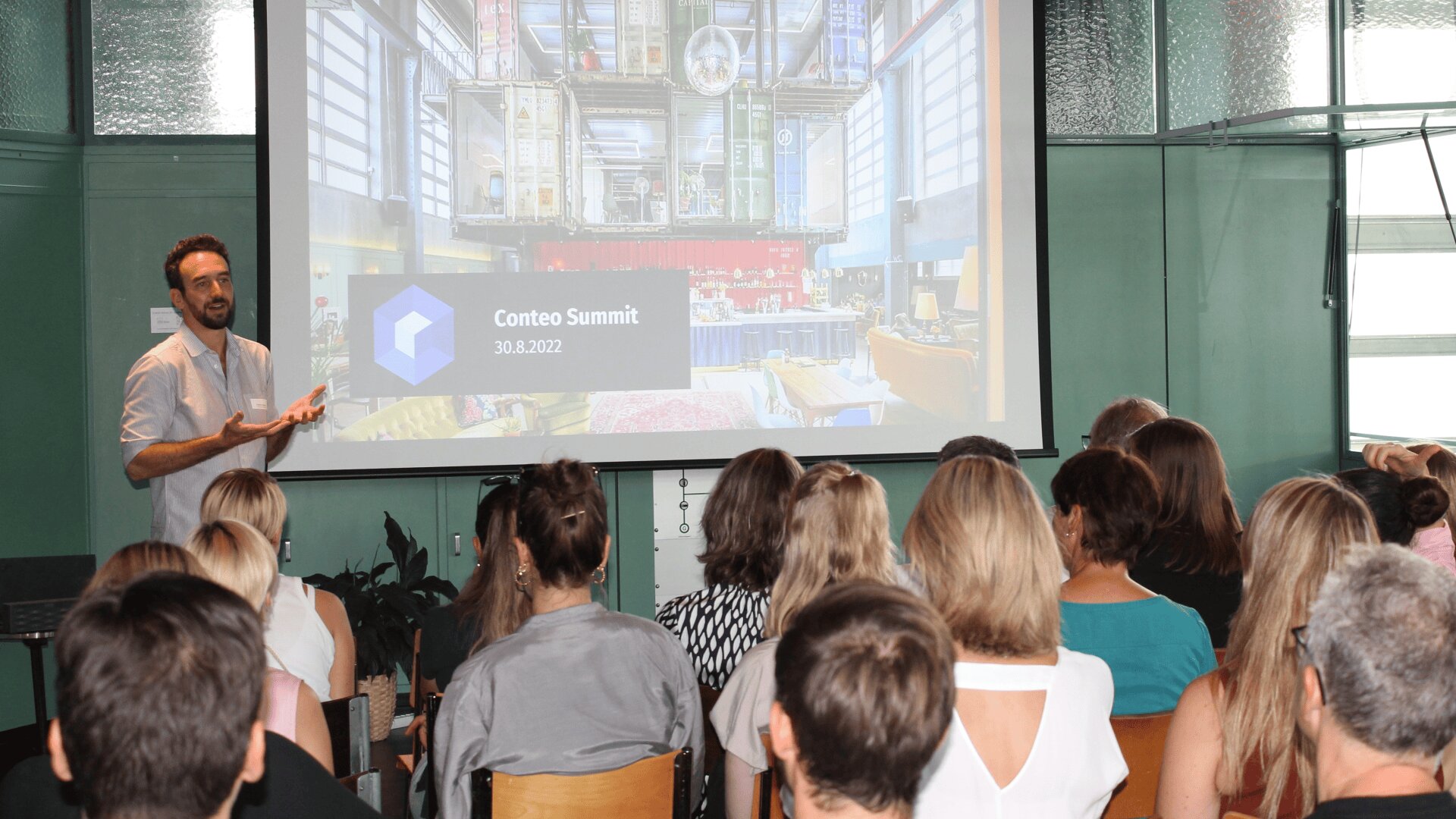 Ein Mann steht vor einem sitzenden Publikum und präsentiert eine Folie mit dem Titel „Conteo Summit 30.8.2022“ mit einem farbenfrohen Innenraumbild, das auf einem großen Bildschirm angezeigt wird.