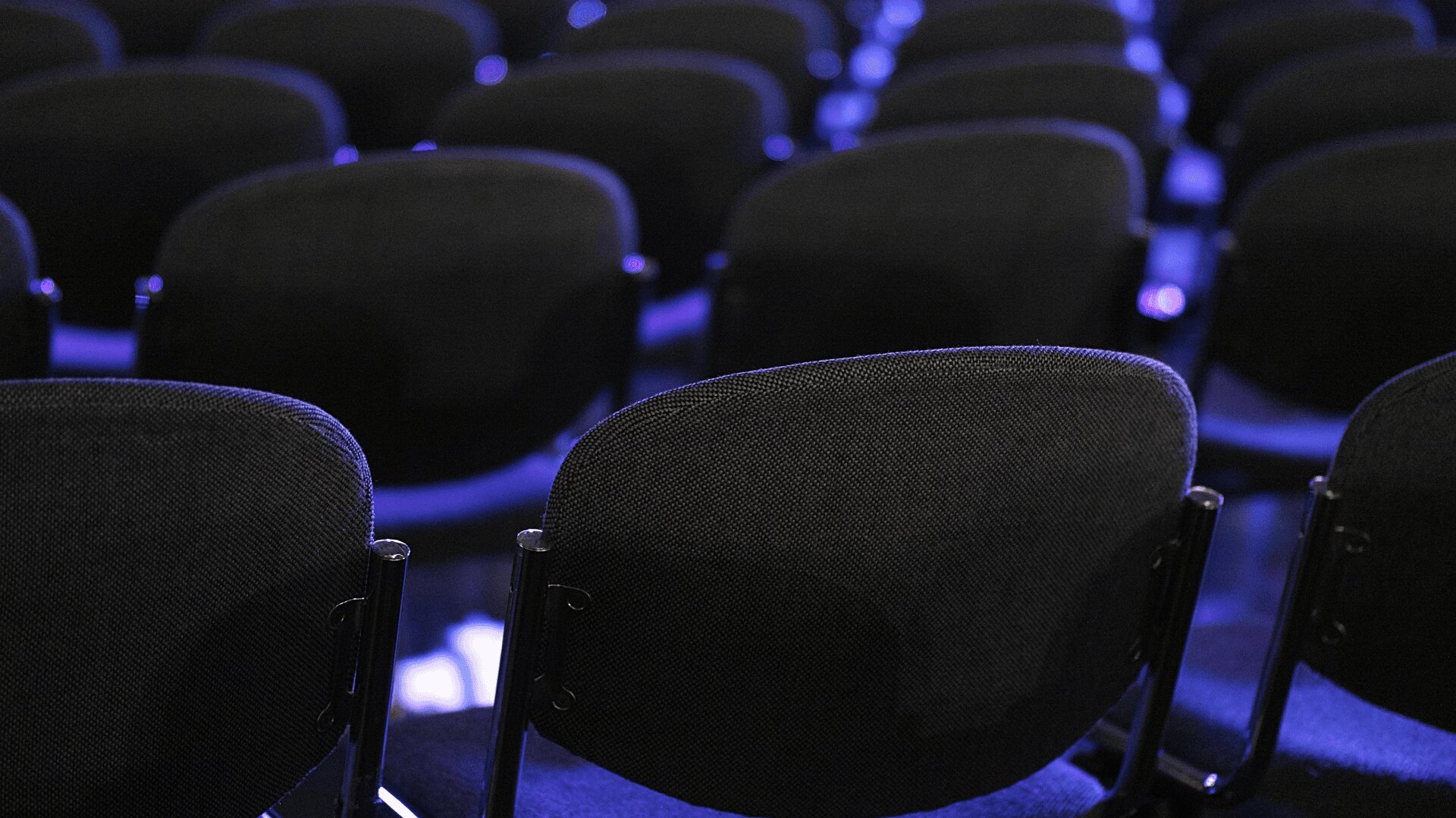 Dicht aneinander gereiht stehen Reihen leerer schwarzer Stühle, die von sanftem blauem und violettem Licht erhellt werden und den Eindruck erwecken, als sei der Veranstaltungsort für eine Veranstaltung oder Präsentation vorbereitet worden.