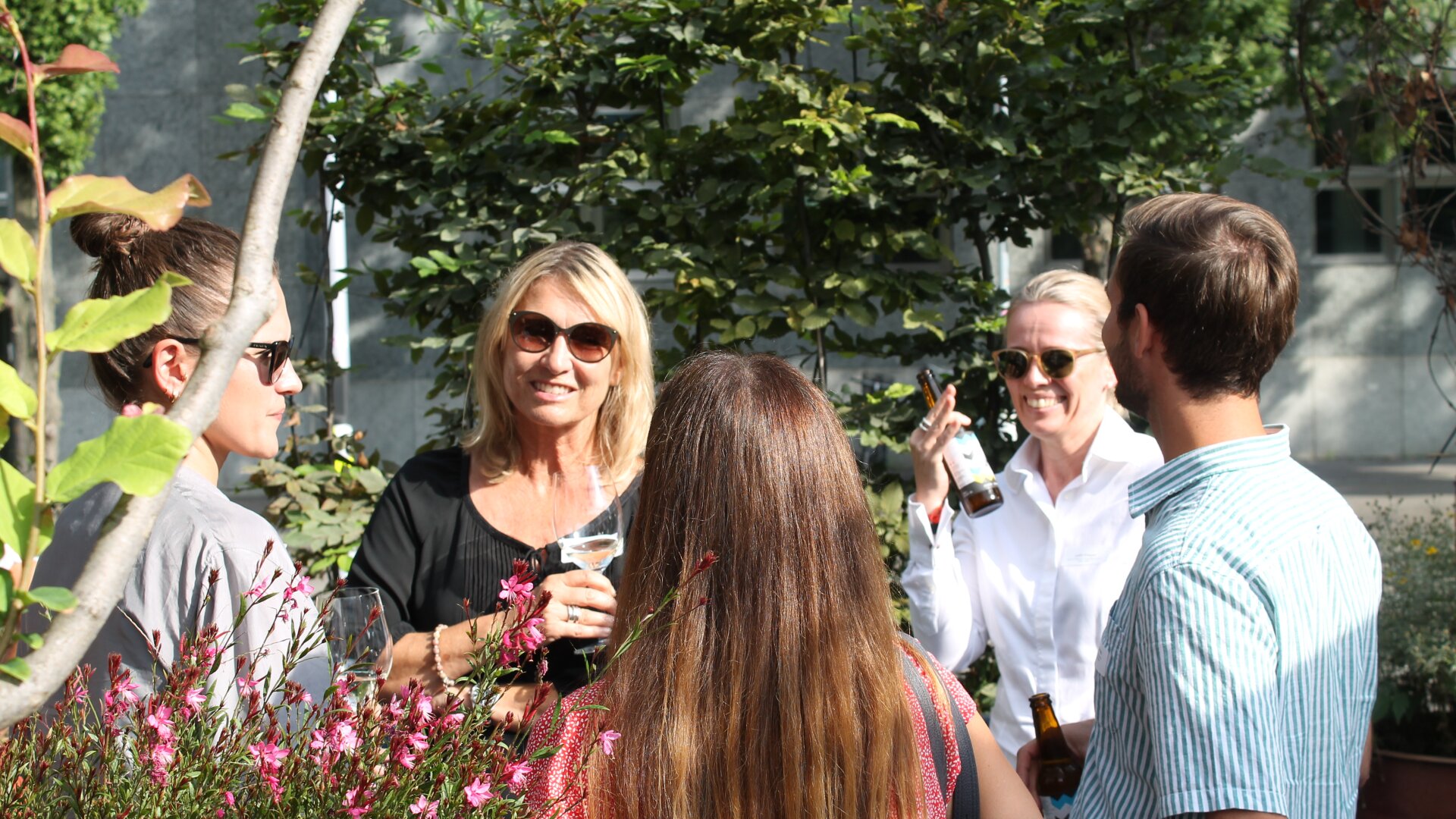 Fünf Erwachsene stehen draußen in einem Garten, unterhalten sich und lächeln, während sie Getränke in den Händen halten. Üppige grüne Bäume und rosa Blumen umgeben sie, und das Sonnenlicht sorgt für eine fröhliche Atmosphäre.