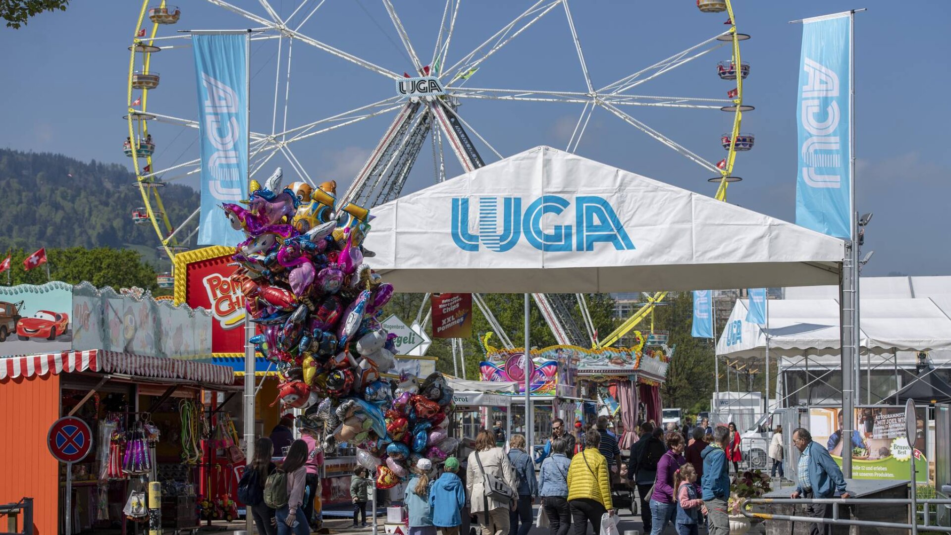 Menschen gehen auf einem Jahrmarkt unter einem weißen Zelt mit der Aufschrift „LUGA“ hindurch. Ein Riesenrad, Buden, Ballons und bunte Stände sind zu sehen, im Hintergrund blauer Himmel und Hügel.