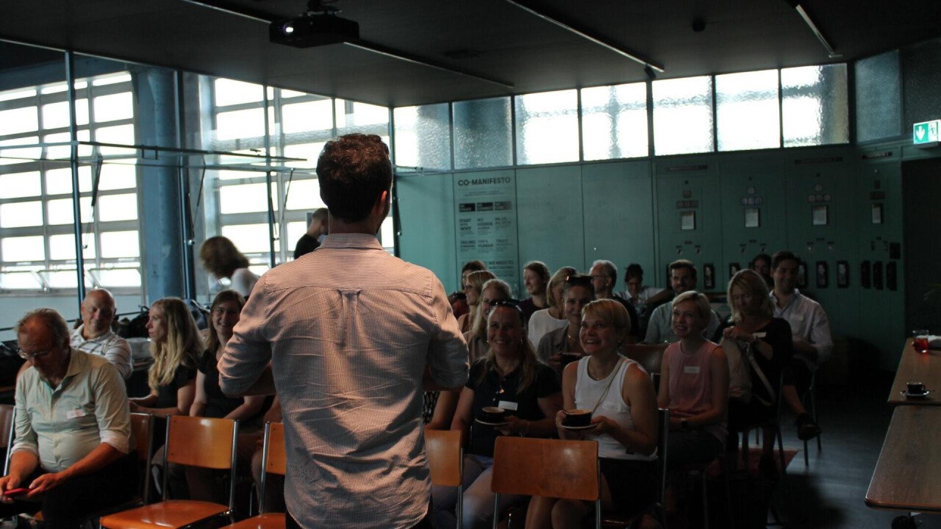A man stands and speaks to a seated audience in a modern, sunlit room with glass walls. The audience appears attentive and engaged, with some people smiling.