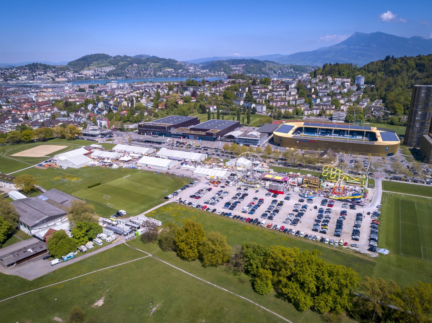 Luftaufnahme eines großen Jahrmarkts mit Fahrgeschäften, einem mit Autos gefüllten Parkplatz, Sportplätzen und einer Stadt mit Hügeln und Bergen im Hintergrund unter einem klaren blauen Himmel.