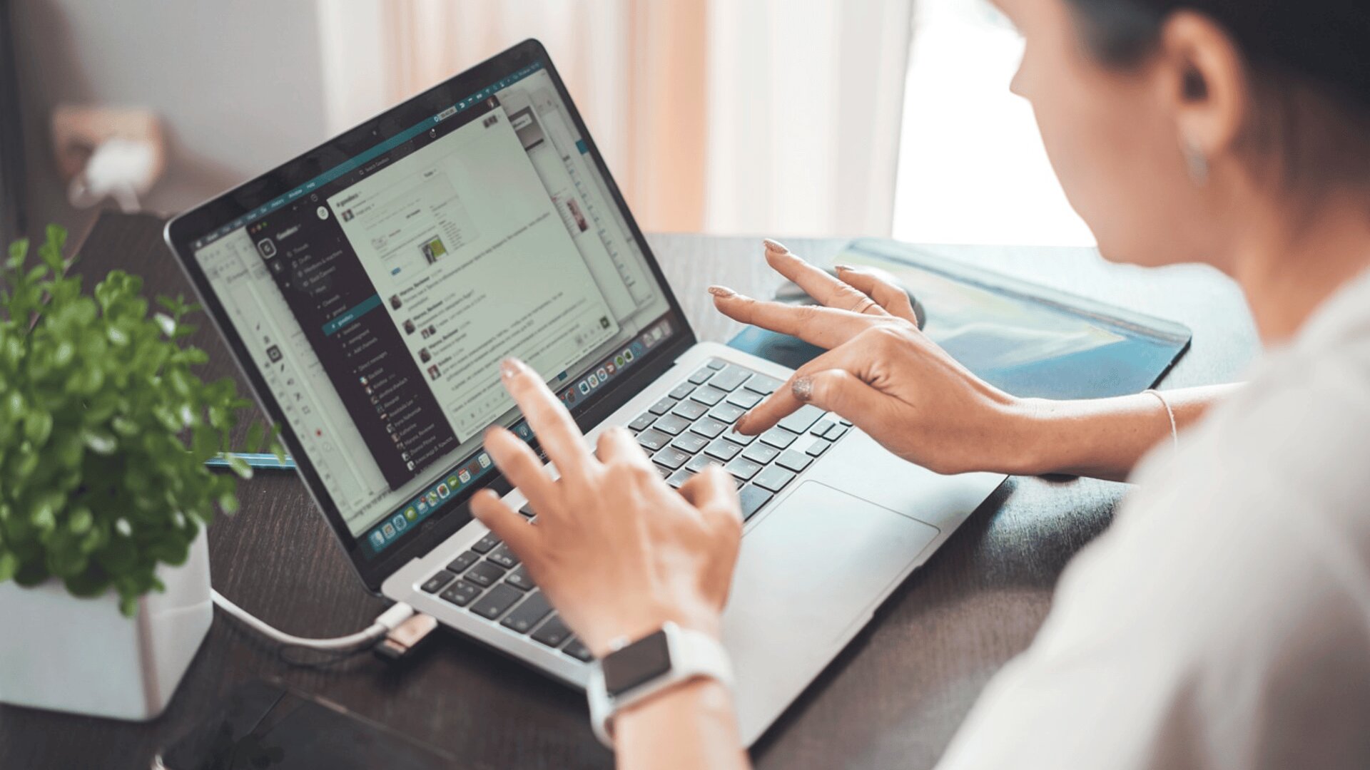 Eine Person sitzt an einem Schreibtisch und nutzt einen Laptop. Auf dem Bildschirm wird eine Messaging-App angezeigt. Eine Topfpflanze und ein Ordner stehen daneben. Die Person trägt eine Smartwatch. Der Arbeitsplatz ist gut beleuchtet.