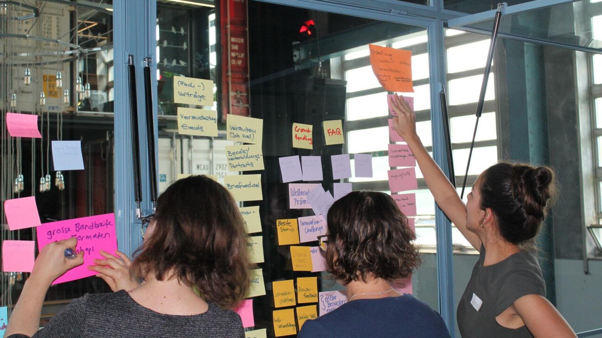 Three people stand in front of a glass wall covered in colorful sticky notes, writing and adding new notes, appearing to collaborate and brainstorm in a bright, modern office space.
