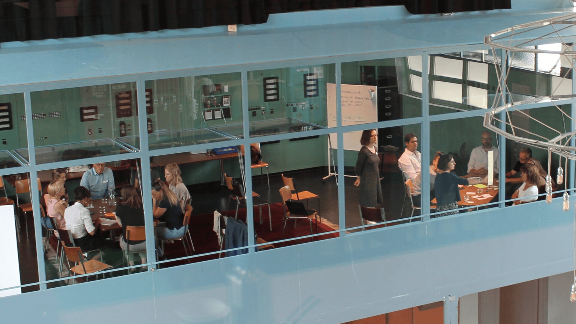 In einem verglasten Besprechungsraum sitzen und stehen Menschen an Tischen und diskutieren und arbeiten gemeinsam. Der Raum verfügt über große Fenster und eine moderne Einrichtung.