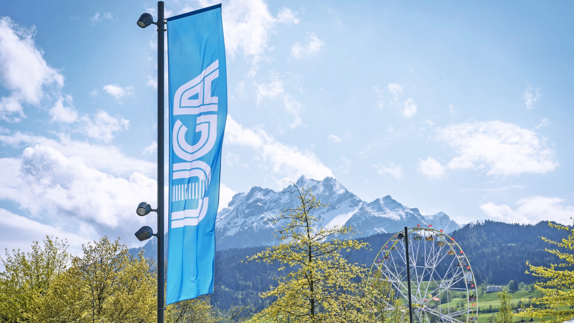 Eine blaue Flagge mit der Aufschrift „LUGA“ steht neben Bäumen, im Hintergrund sind unter einem teilweise bewölkten Himmel ein Riesenrad und schneebedeckte Berge zu sehen.