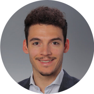 A young man with short curly brown hair, fair skin and a trimmed goatee smiles at the camera. He is wearing a light-colored shirt and a dark blazer against a plain gray background.